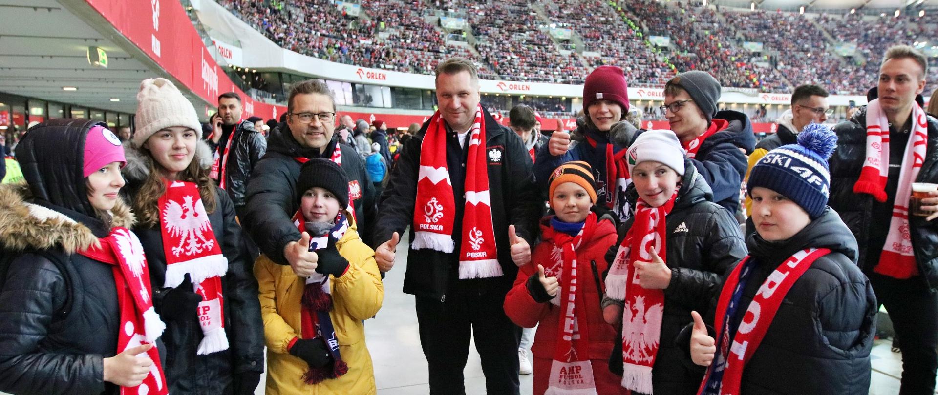 Minister Czarnek w biało-czerwonym szaliku stoi na białej podłodze, wokół niego dzieci w takich samych szalikach, w tle trybuny stadionu.
