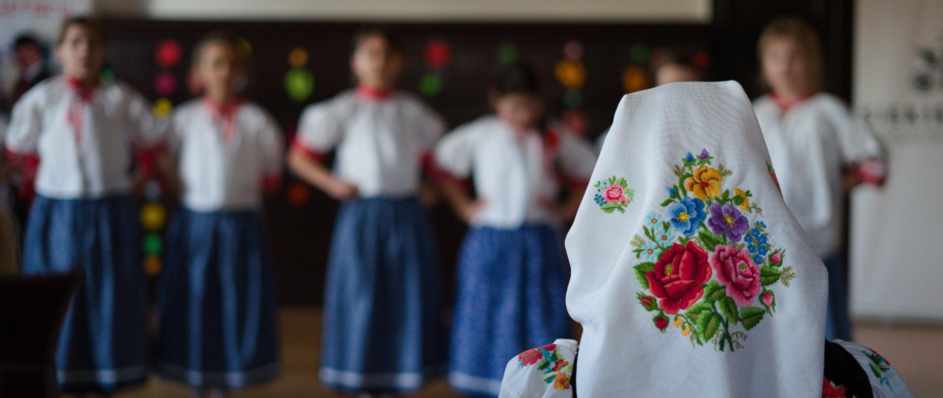 Zdjęcie, na którym na pierwszym planie widać tył głowy kobiety ubranej w tradycyjną chustę z wzorem łowickim. W tle można dostrzec lekko rozmazane dziewczynki w podobnym wieku (ok 10 lat) podczas występu wokalnego. Wszystkie stoją w jednym rzędzie obok siebie. Mają ręce oparte na biodrach. Są ubrane w białe haftowane bluzki z czerwonymi wstawkami w okolicy szyi i ramion. Mają niebieskie spódnice do kostek w mały, drobny wzór białych kwiatów. Na nogach założone mają baletki. Stroje imitują beskidzki strój ludowy.