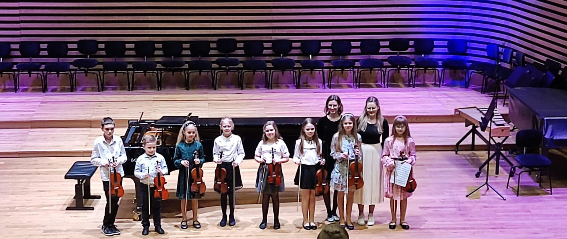 Ośmioro dzieci trzymających przed sobą skrzypce stoi w rzędzie, zaraz za nimi po prawej stronie stoją dwi kobiety. Za nimi stoi fortepian i z tyły ustawione pod ścianą krzesła.