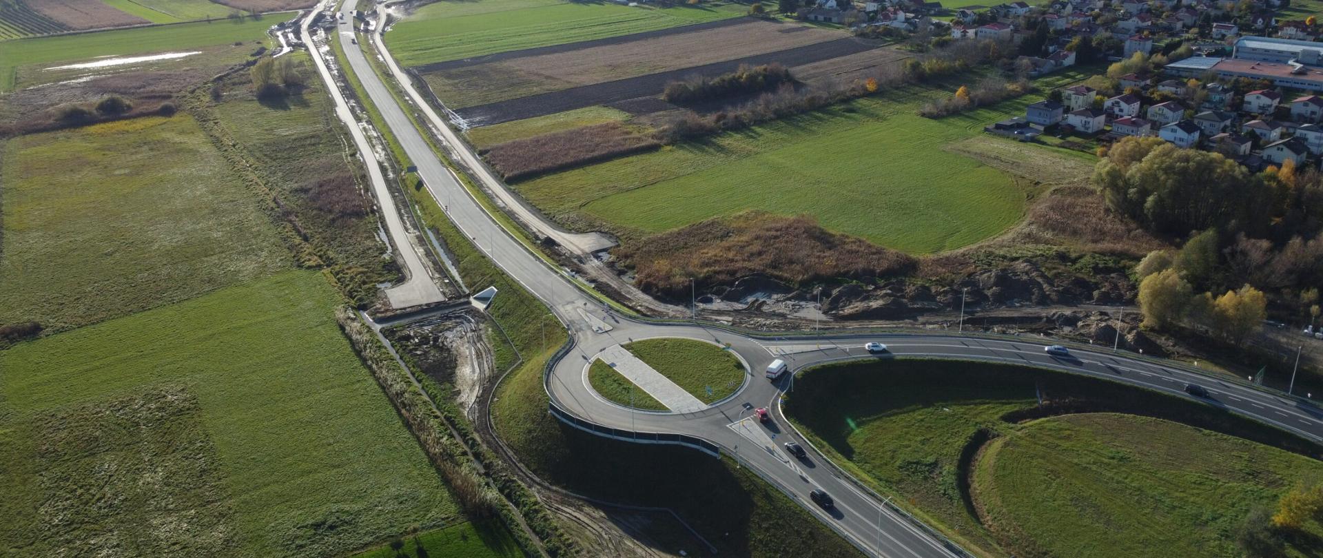 DK9 obwodnica Ostrowca Świętokrzyskiego - zdjęcie z drona, jednojezdniowa droga asfaltowa wśród pól, po obu stronach wąskie drogi dojazdowe, na pierwszym planie rondo trójwlotowe, w oddali zabudowania 