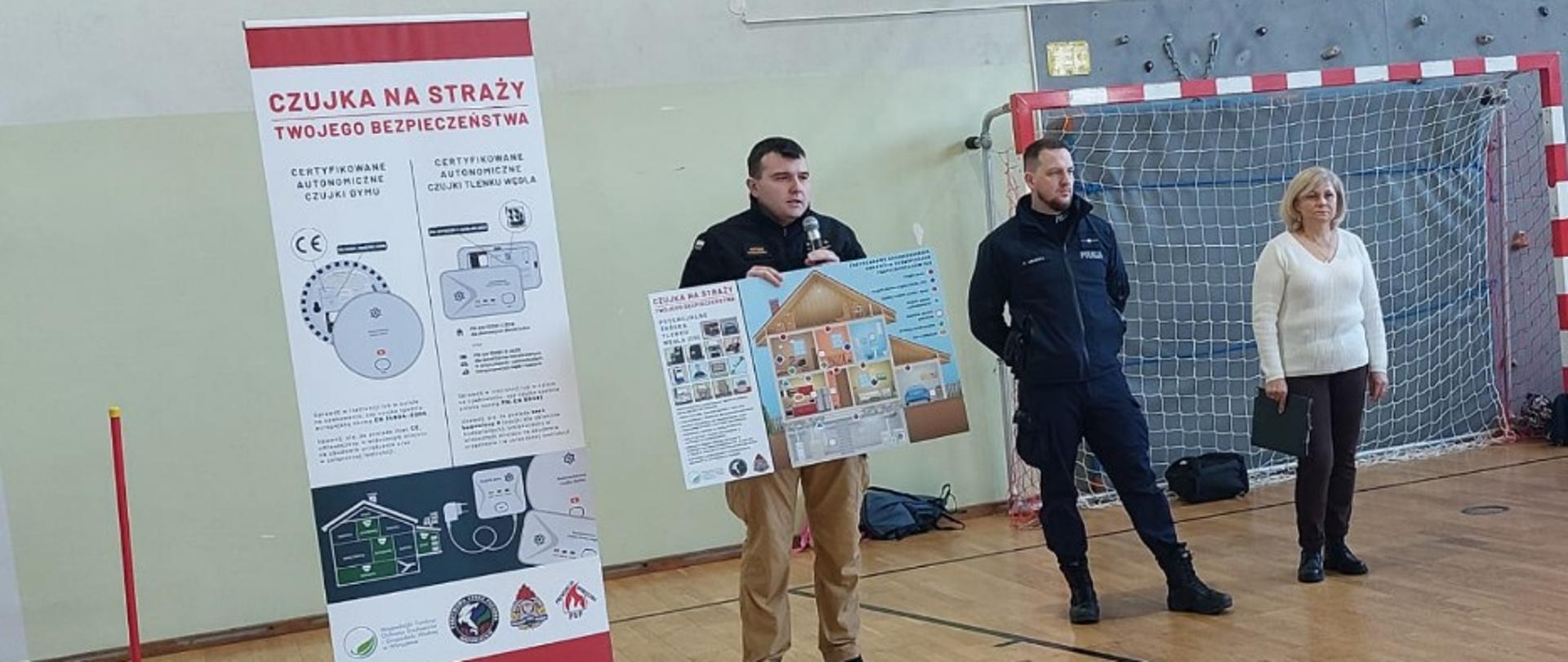Zdjęcie przedstawia funkcjonariusz PSP prezentującego materiały na temat akcji "Czujka na Straży Twojego bezpieczeństwa", obok funkcjonariusz Policji i nauczyciel. Zdjęcie wykonane na sali gimnastyczne, w tle bramka i ściana wspinaczkowa.