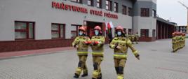 Trzech strażaków, jako poczet flagowy maszeruje do niewidocznego masztu. Z tyłu Strażacy w ubraniach specjalnych ustawieni w dwóch szeregach. W tle budynek Komendy Powiatowej PSP w Płońsku. 
