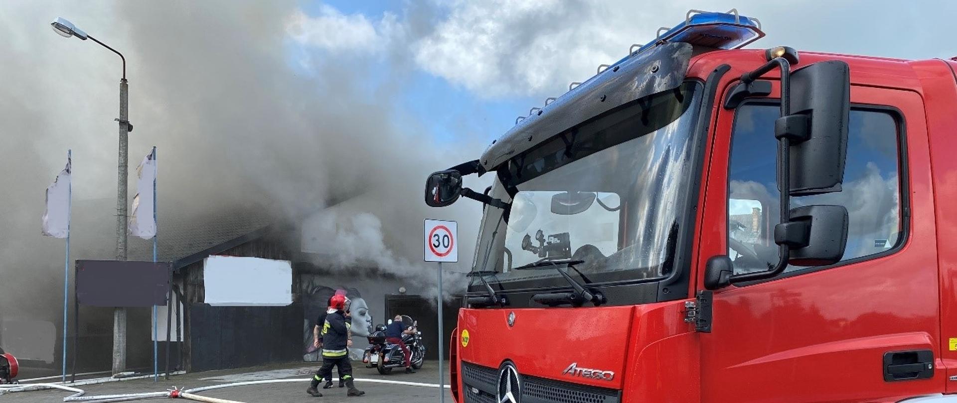 Pożar budynek warsztatu samochodowego. Widoczne kłęby dymu nad budynkiem. Na pierwszym planie samochód ratowniczo - gaśniczy Komendy Powiatowej Państwowej Straży Pożarnej w Lęborku
