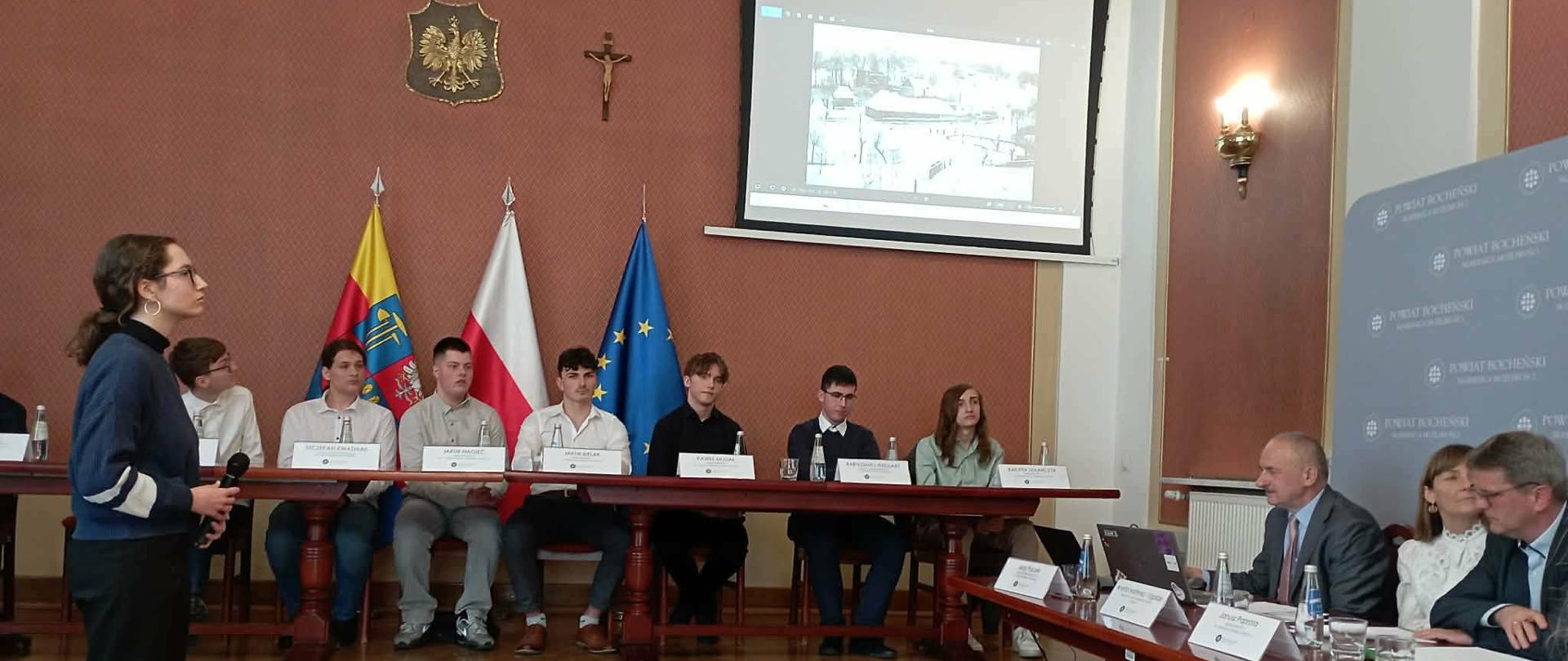 W centrum sali znajduje się długi stół, przy którym siedzi kilkanaście osób, przed nimi stoją wizytówki z imionami i nazwiskami oraz butelki wody.
Na pierwszym planie po prawej stronie widać kolejny stół z trzema osobami, członkami komisji.
Po lewej stronie kadru widać osobę stojącą z mikrofonem, zwróconą w stronę komisji. W tle, za uczestnikami, widoczne są flagi: flaga Polski, flaga Unii Europejskiej oraz flaga samorządowa.
Na ścianie znajduje się godło Polski oraz krzyż.
Po prawej stronie ustawiona jest ścianka z powtarzającym się napisem „Powiat Bocheński”.
Nad uczestnikami wisi ekran projekcyjny, na którym wyświetlane jest zdjęcie (widok zabudowań/miasta).
