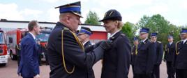 Komendant wojewódzki PSP w Łodzi nadaje oznaczenie dla kierownika sekcji kwatermistrzowskiej st. kpt. Barbary Błaszczyk, w tle funkcjonariusze oczekujący na nominacje, po lewej stronie komendanta wojewódzkiego wicestarosta łowicki gratuluje komendantowi powiatowemu mł. bryg. Arkadiuszowi Makowskiemu, w tle budynek garażowy komendy i samochody pożarnicze. 