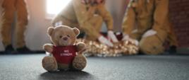 Miś strażak na podłodze, w tle strażacy w trakcie praktycznego egzaminu kwalifikowanej pierwszej pomocy