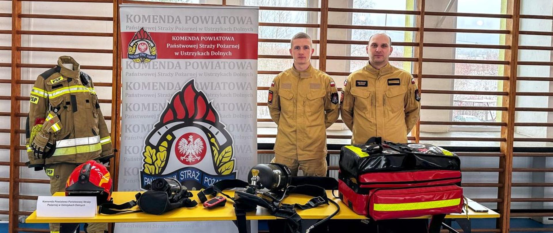 Na zdjęciu widać dwóch funkcjonariuszy straży pożarnej. Zdjęcie wykonane jest na hali sportowej gdzie znajduje się stoisko Państwowej Straży Pożarnej. 