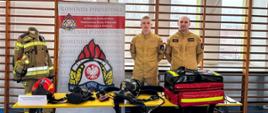 Na zdjęciu widać dwóch funkcjonariuszy straży pożarnej. Zdjęcie wykonane jest na hali sportowej gdzie znajduje się stoisko Państwowej Straży Pożarnej. 