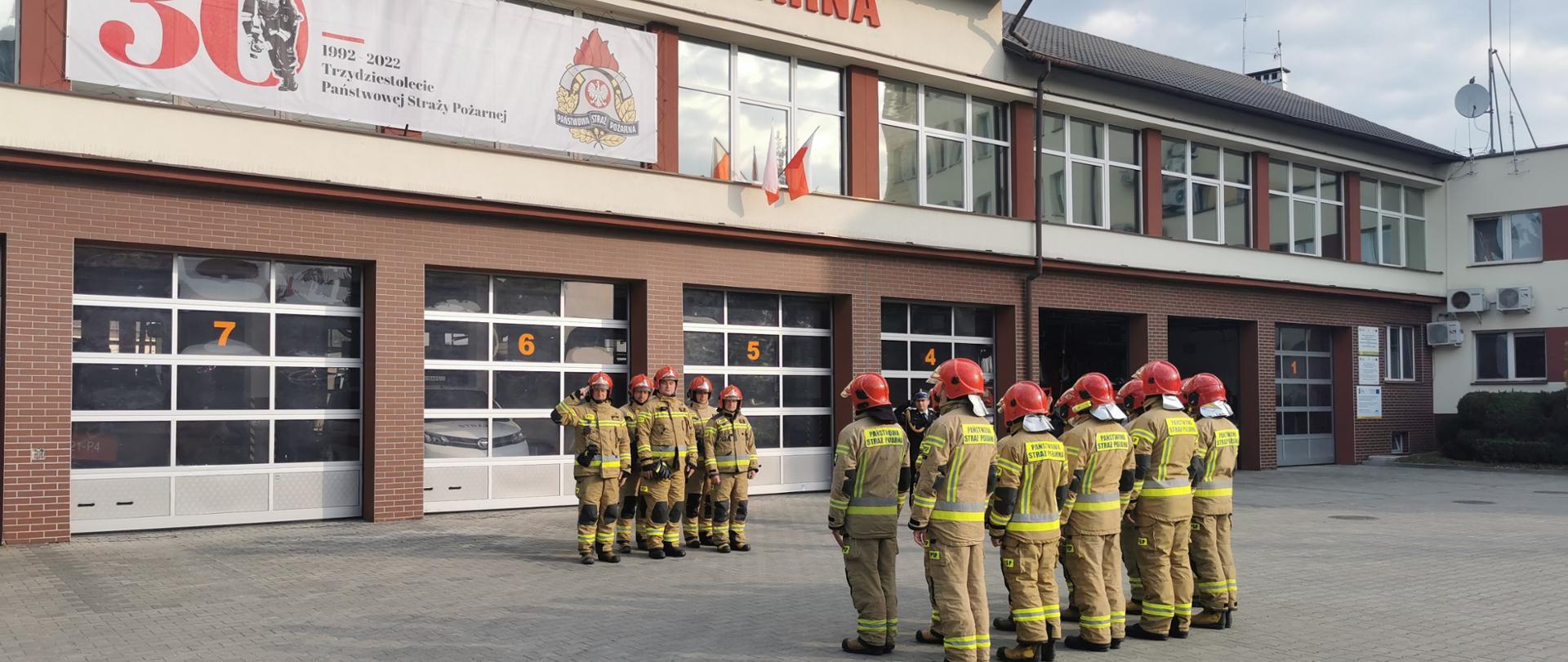 Zdjęcie przedstawia strażaków w umundurowaniu bojowym dwóch zmian służbowych stojących naprzeciwko siebie na tle garażu Jednostki Ratowniczo-Gaśniczej Nr 1 w Rzeszowie.