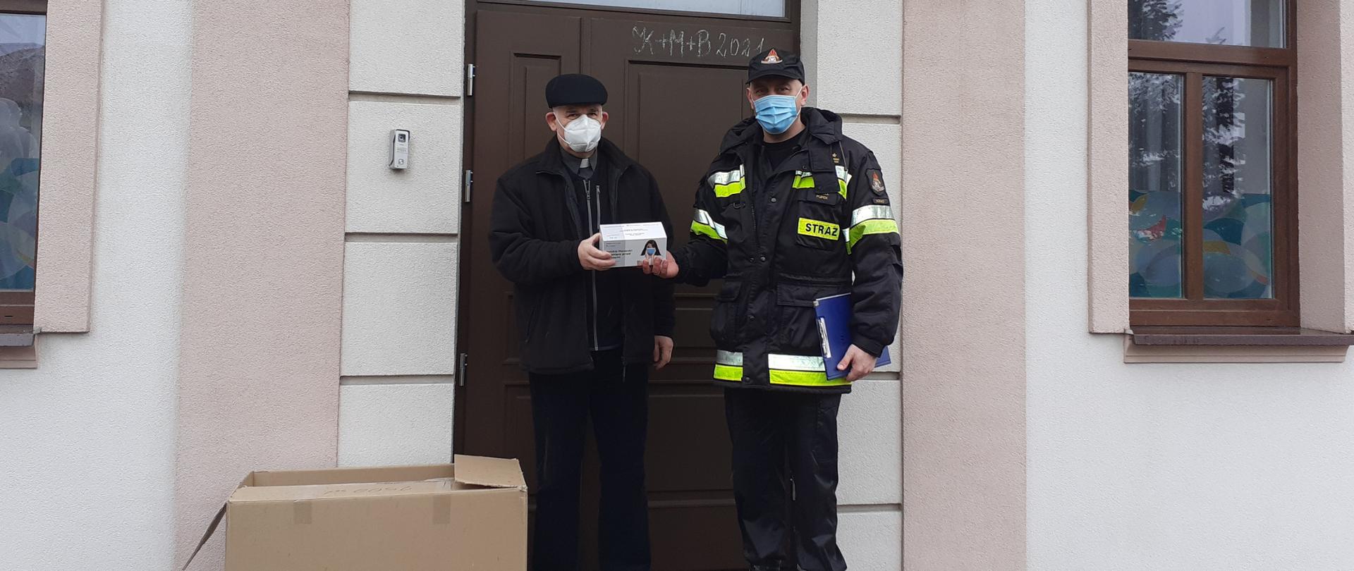 Widać strażaka i księdza. Ksiądz trzyma w ręku paczkę maseczek. Na dalszym planie widać część budynku. Po lewej stronie księdza stoi karton, za nim znajdują się drzwi do budynku.