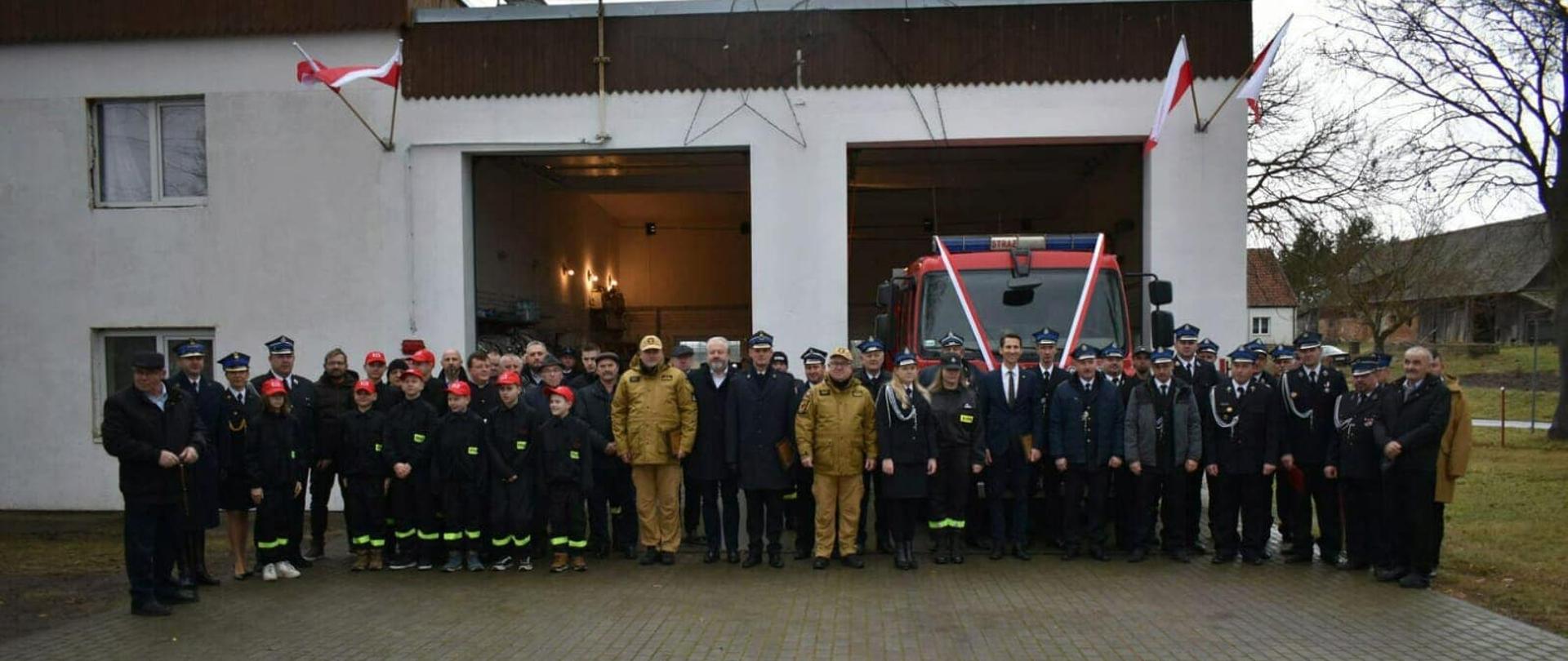 Uczestnicy uroczystej zbiórki pozują do zdjęcia na tle remizy.