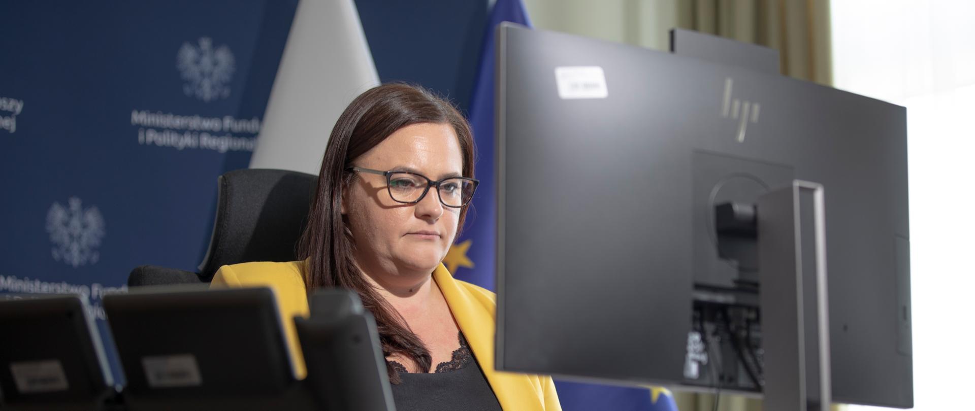 Wiceminister Małgorzata Jarosińska-Jedynak przed monitorem. Za nią ścianka ministerialna i flagi PL i UE.