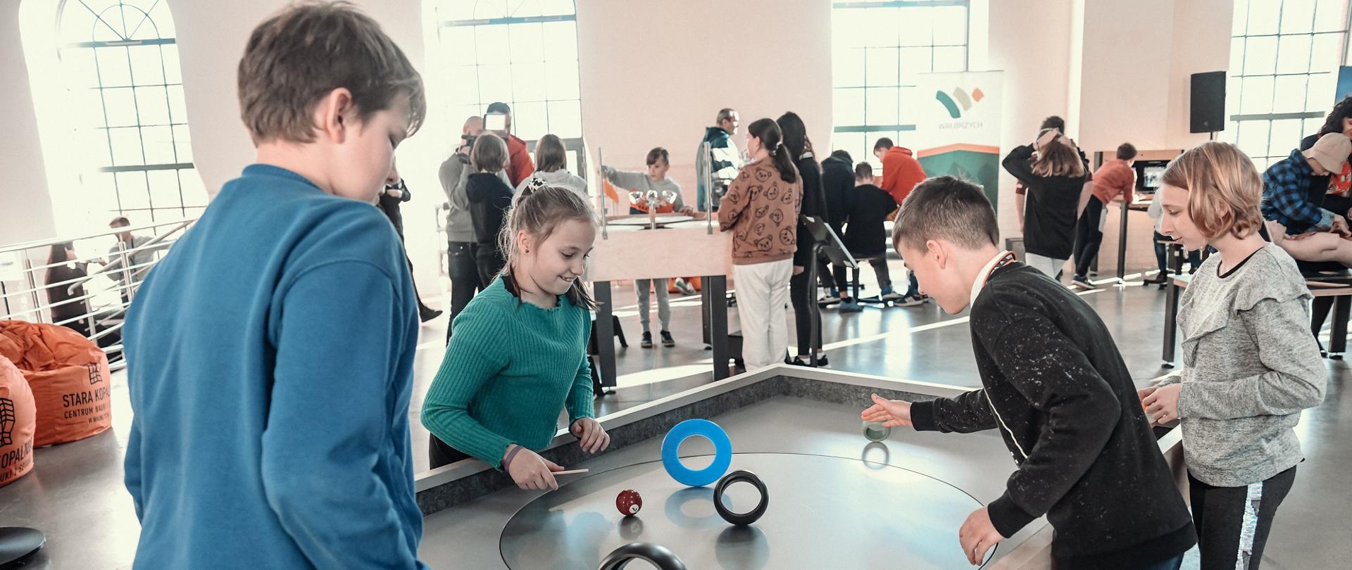 Kilkoro dzieci stoi przy prostokątnym stole, puszczają po nim kolorowe kółka.