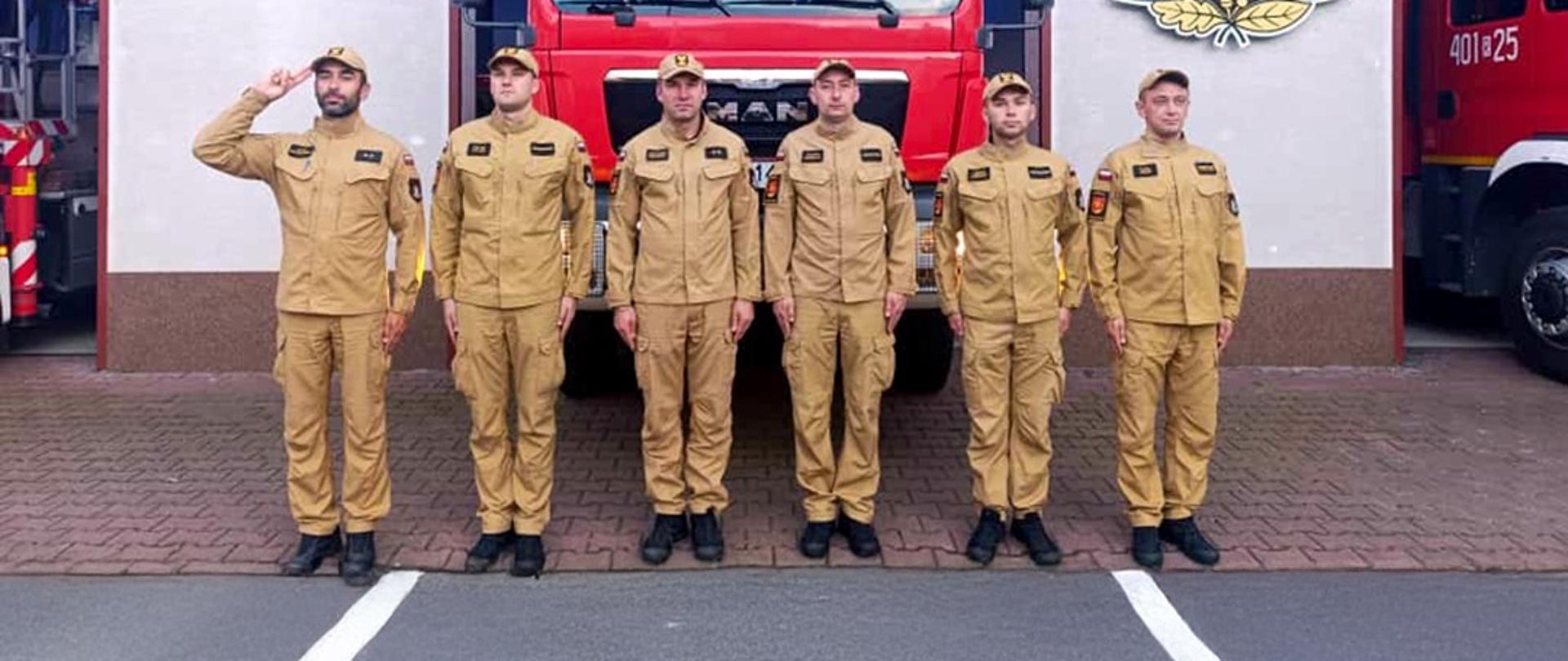 Zdjęcie przedstawia sześciu funkcjonariuszy Państwowej Straży Pożarnej przed garażem JRG. Z lewej strony dowódca oddaje honor a reszta strażaków stoi na baczność. Za nimi widać samochód gaśniczy z włączonymi światłami błyskowymi.