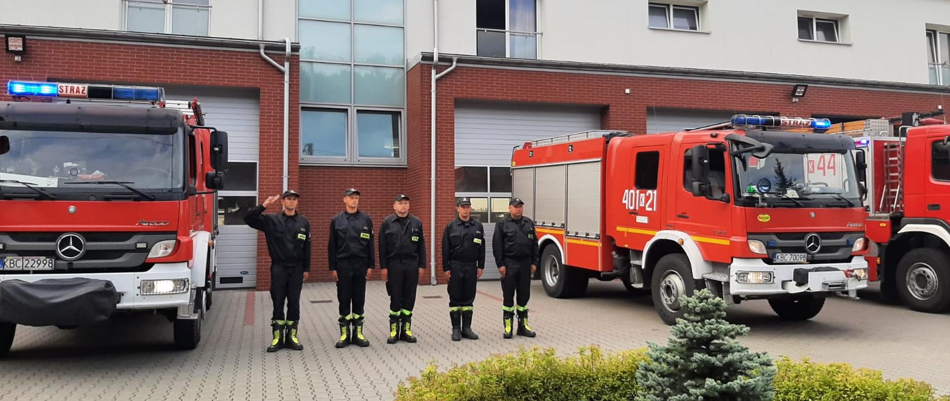 Na zdjęciu widać na kostce brukowej stoją dwa czerwone ciężarowe pojazdy pożarnicze oraz drzwi cześć zabudowy trzeciego pojazdu pożarniczego w kolorze czerwonym . W dwóch pojadać widać włączone światła drogowe i światło błyskowe niebieskie. Między dwoma pojazdami pożarniczymi marki mercedes stoją w szeregu pięciu strażaków ubranych w czarne mundury koszarowe z odblaskowymi paskami i czapeczkami na głowach w kolorze czarnym. Pierwszy po lewej strażak salutuje dwoma palcami do daszku czapki. Za samochodami ciężarowymi stoi terenowy samochód pożarniczy. Po lewej stronie znajduje się żywopłot a na środku drzewo iglaste.. Za pojazdami pożarniczymi znajduje się budynek Komendy Powiatowej w kolorze białym . Na dole znajdują się bramy garażowe w kolorze białym i szybami przez środek drzwi” Przed pojazdami jest wysepka ogrodzona żywopłotem na środku znajduje się małe drzewo iglaste.