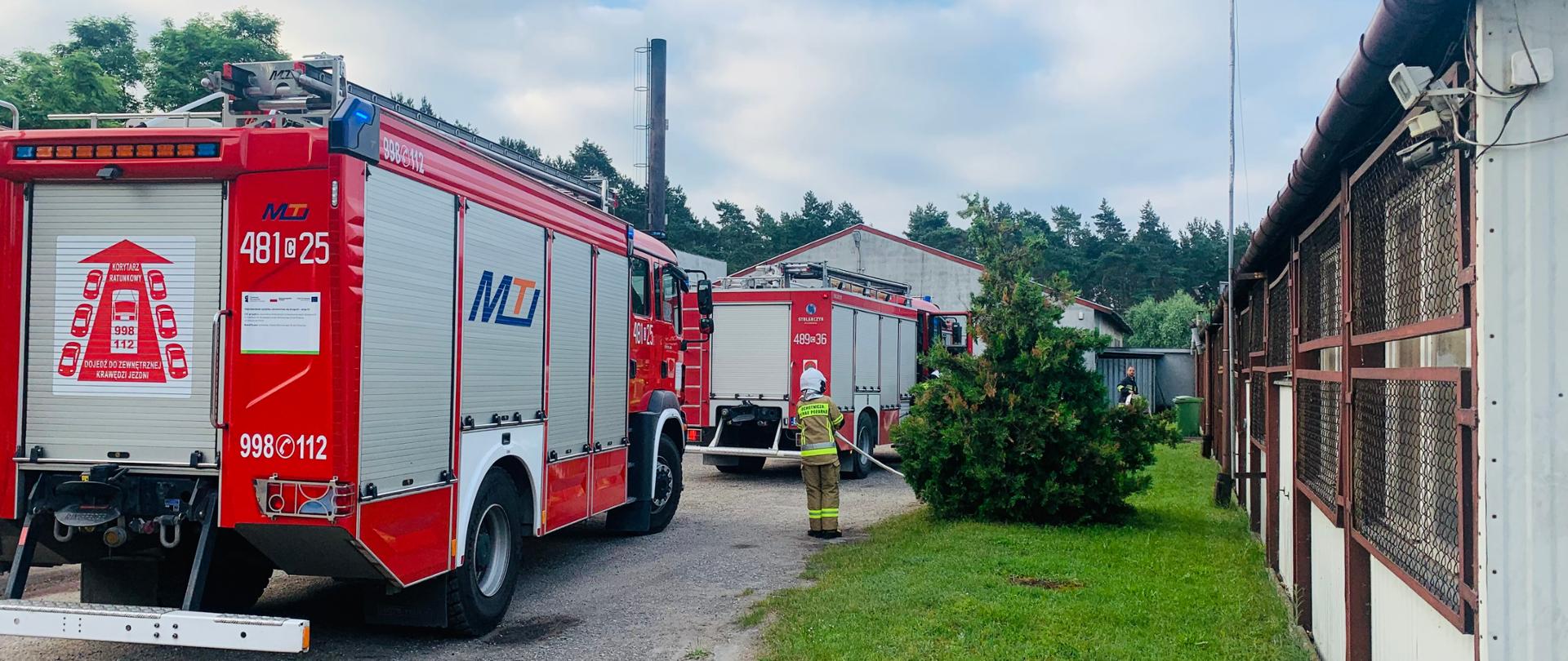 Po lewej stronie stoją 2 pojazdy pożarnicze. Przed nimi i po prawej stronie budynki tartaku i lakierni. Przy samochodzie stoi strażak