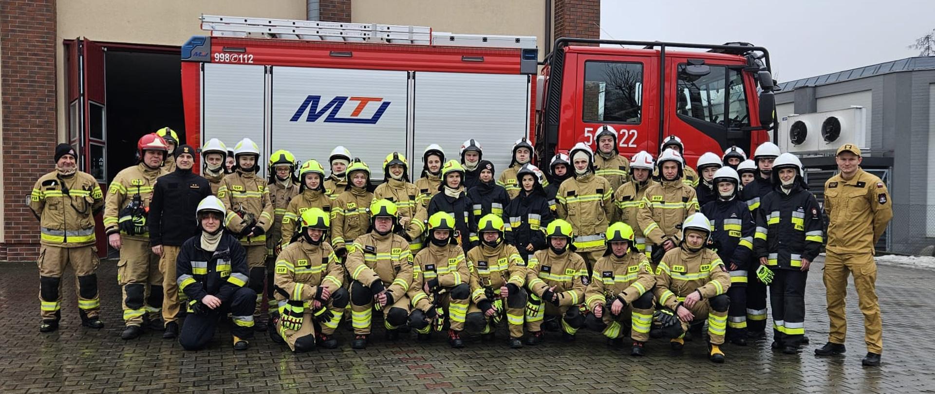 Na zdjęciu kursanci w ubraniach specjalnych pozują przed wozem strażackim. W tle budynek strażnicy.