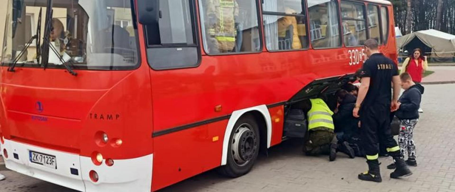 Centralny punkt zdjęcia stanowi czerwony strażacki autobus. Przy autobusie stoi grupka ludzi i pakuje torby do bocznych luków. W tle widać las i namioty.