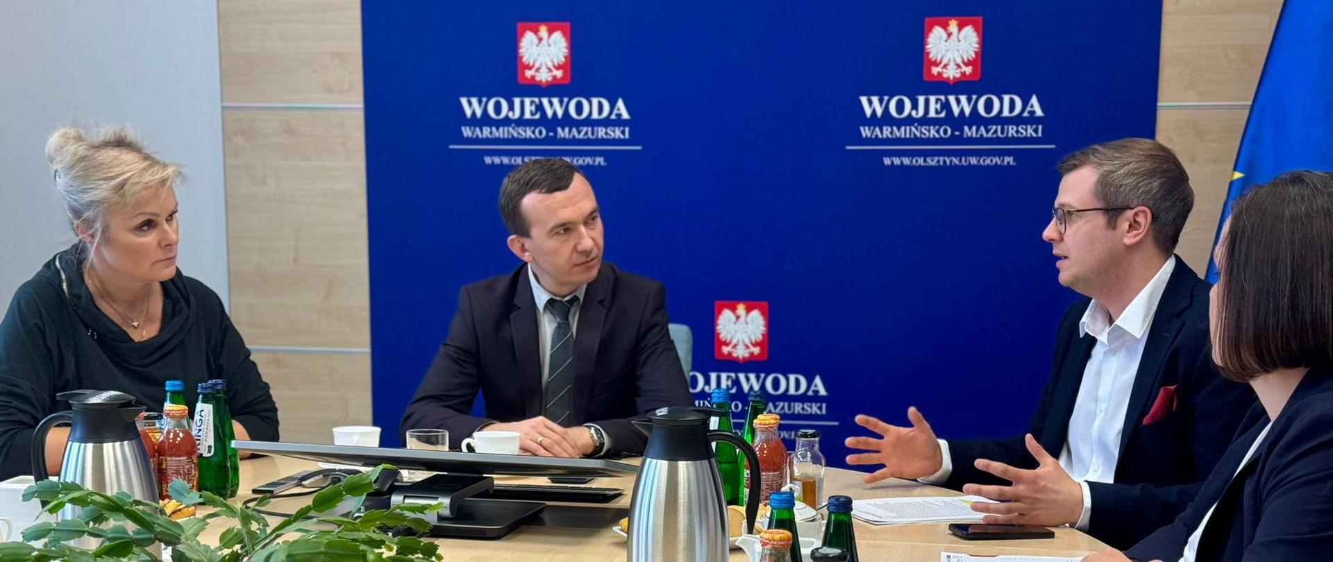 4 osoby rozmawiają przy stole konferencyjnym na tle ścianki wojewoda warmińsko-mazurski 