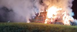Widok z przodu na płonący stóg balotów słomy. Stóg w częściach bocznych i środkowej objęty płomieniami. 