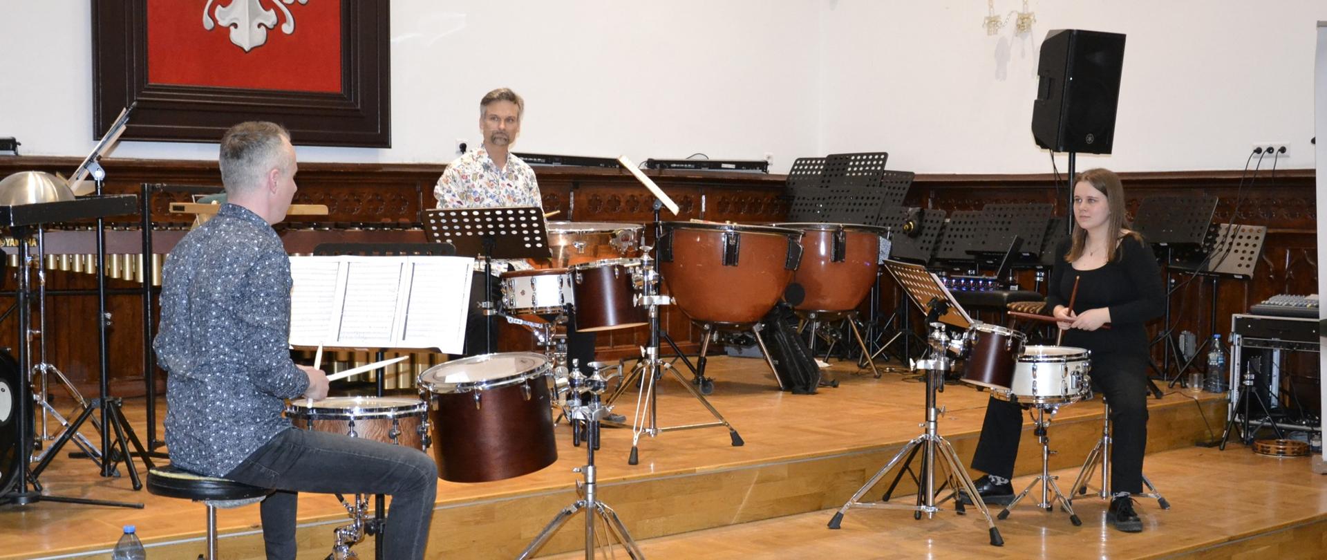 Trzech perkusistów gra na instrumentach perkusyjnych na scenie podczas warsztatów i koncertu dra Piotra Szulca w sali koncertowej szkoły muzycznej.