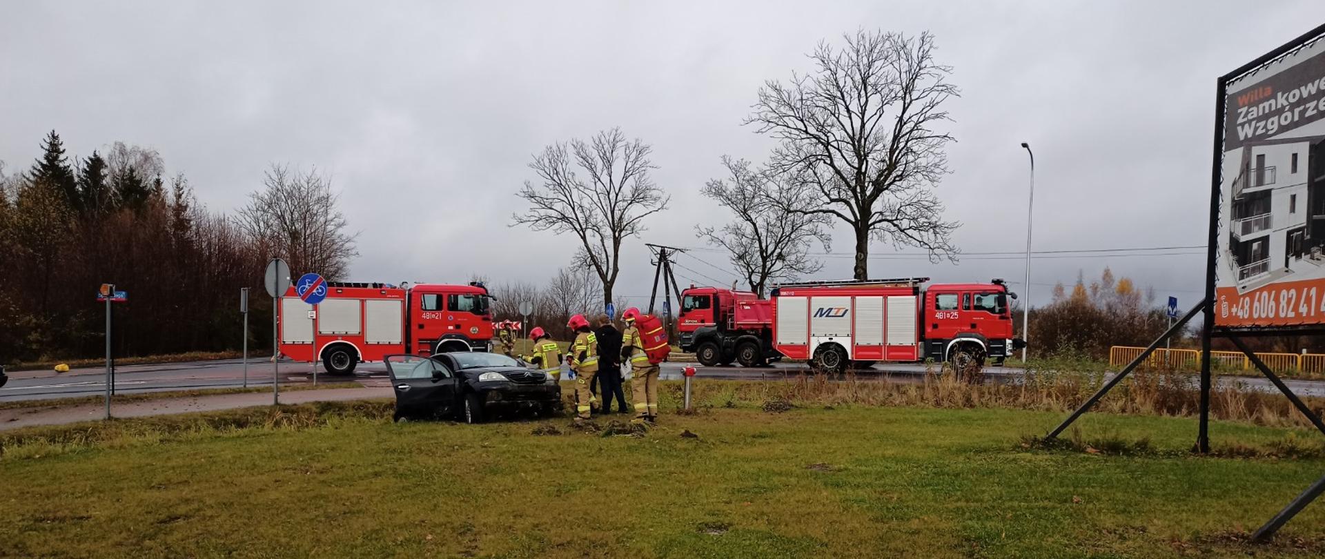 Zdjęcie przedstawia uszkodzony samochód znajdujący się częściowo w rowie poza drogą wojewódzką. Obok niego znajdują się strażacy.
