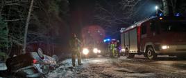 Jezdnia oraz pobocze drogi pokryte śniegiem. W przydrożnym rowie leży samochód w pozycji na dachu. Na jezdni pojazdy straży pożarnej z włączonymi sygnałami świetlnymi. Obok pojazdu stoi strażak w ubraniu bojowym. Zdarzenie ma miejsce w porze nocnej.