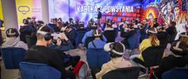 W dużej sali z kolorowymi ścianami z napisem Kartka z powstania siedzi wielu młodych ludzi, na głowach mają gogle do wirtualnej rzeczywistości.