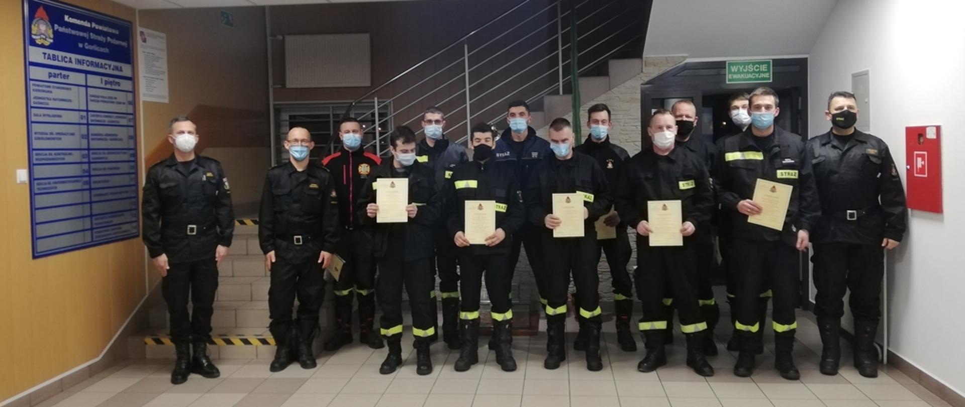 Na zdjęciu strażacy Ochotniczych Straży Pożarnych w ubraniach koszarowych bez nakrycia głowy po ukończonym szkoleniu Naczelników OSP z terenu powiatu gorlickiego oraz komisja egzaminacyjna w ubraniach dowódczo sztabowych koloru czarnego. 