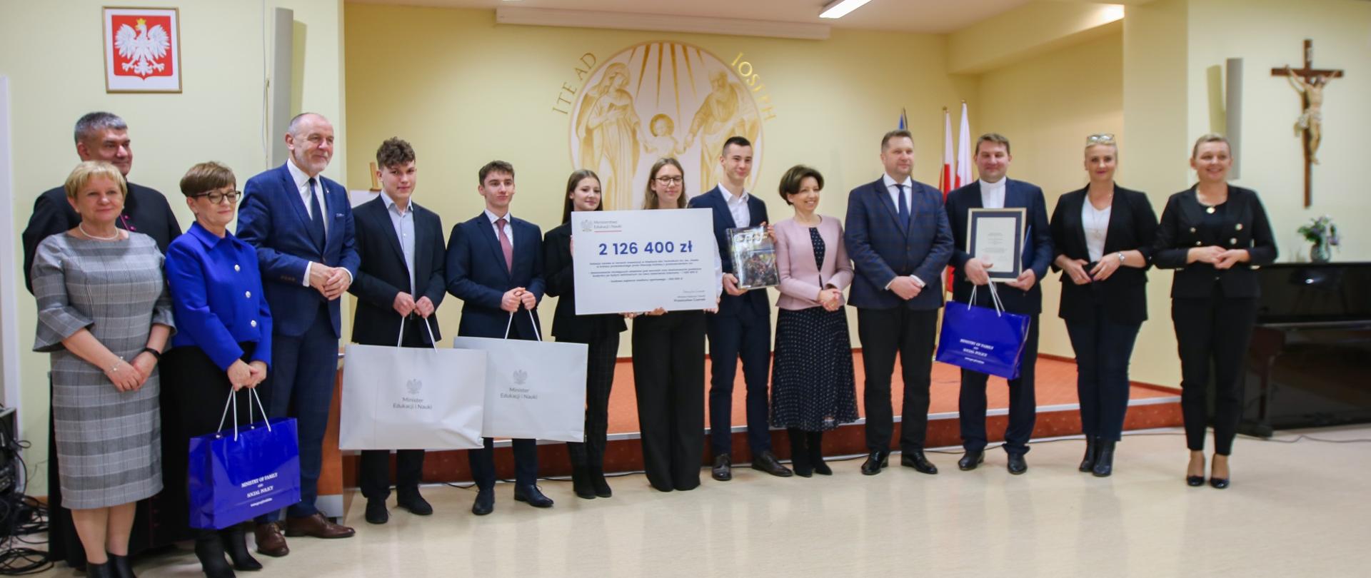 Zdjęcie zbiorowe, w sali stoi duża grupa ludzi, dwie dziewczyny trzymają duży symboliczny czek z napisem 2 126 400 zł.