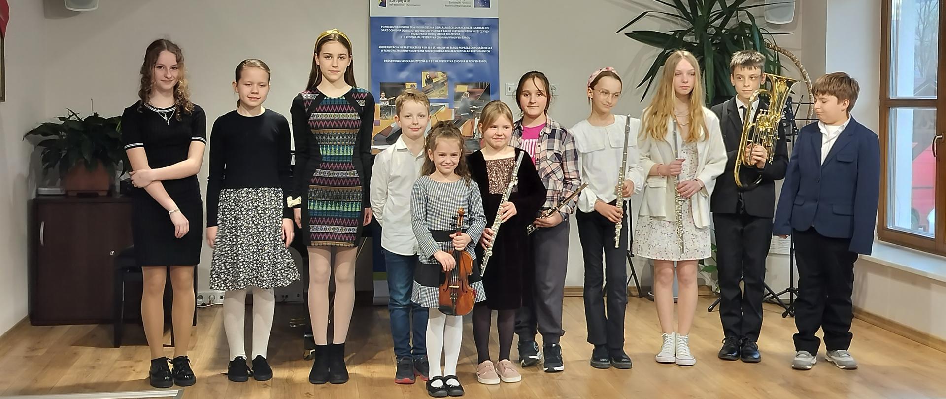 Aula koncertowa filii w Jaworkach. Dziesięcioro dzieci stoi na podeście w rzędzie jedna dziewczynka trzymająca skrzypce lekko z przodu. Trzy dziewczynki trzecia, czwarta i szósta od prawej trzymają flety. Drugi od prawej chłopiec trzyma trąbkę. W tle brązowa szafka na której stoi kwiatek , na środku baner reklamowy otrzymanych środków z Unii Europejskiej, po prawej kwiatek. 
