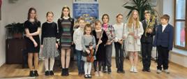 Aula koncertowa filii w Jaworkach. Dziesięcioro dzieci stoi na podeście w rzędzie jedna dziewczynka trzymająca skrzypce lekko z przodu. Trzy dziewczynki trzecia, czwarta i szósta od prawej trzymają flety. Drugi od prawej chłopiec trzyma trąbkę. W tle brązowa szafka na której stoi kwiatek , na środku baner reklamowy otrzymanych środków z Unii Europejskiej, po prawej kwiatek. 
