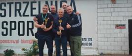 na podium na tle ściany budynku strzelnicy w miejscowości Panaszew stoją reprezentanci komendy powiatowej psp która w zawodach strzeleckich zajęła drugie miejsce, reprezentanci stoją na podium trzymając w rękach medale i puchary za zawody