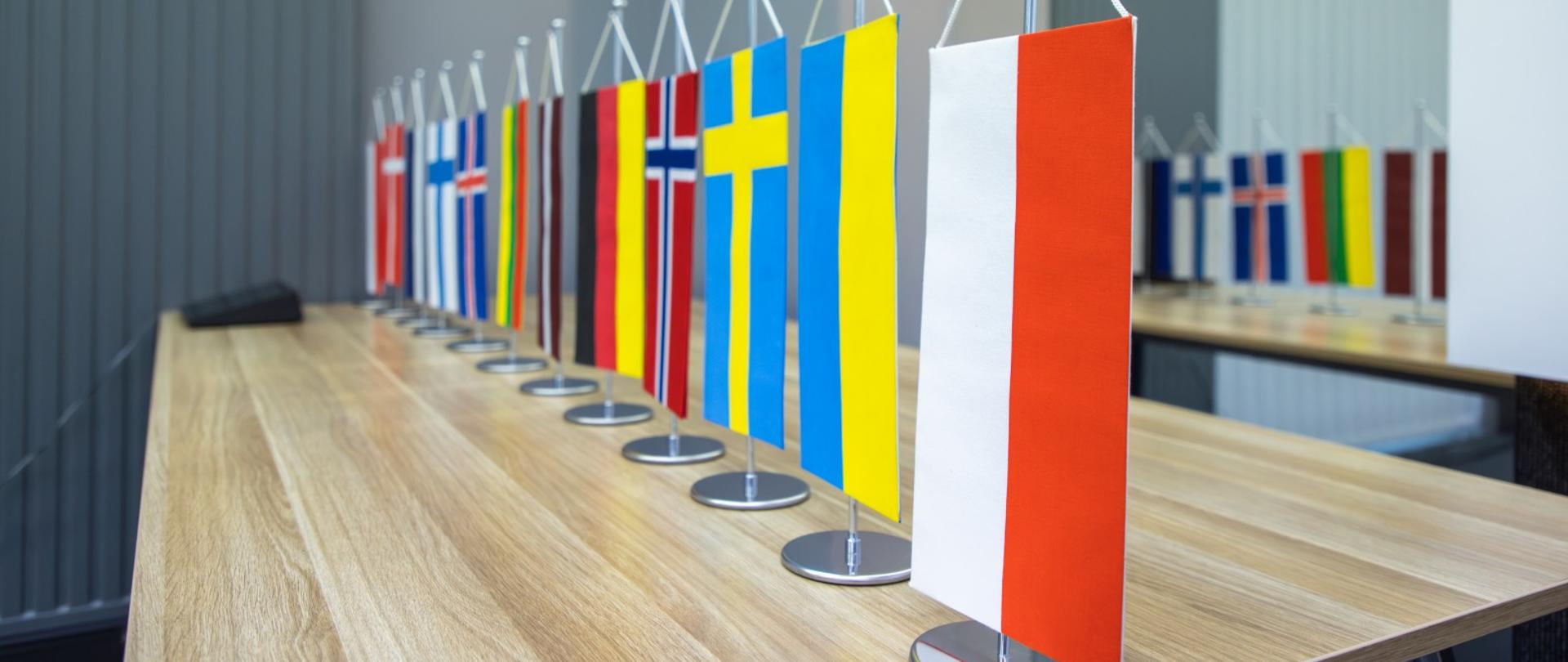 Rząd małych flag państw europejskich ustawionych na stole konferencyjnym. Na pierwszym planie widoczna jest flaga Polski.