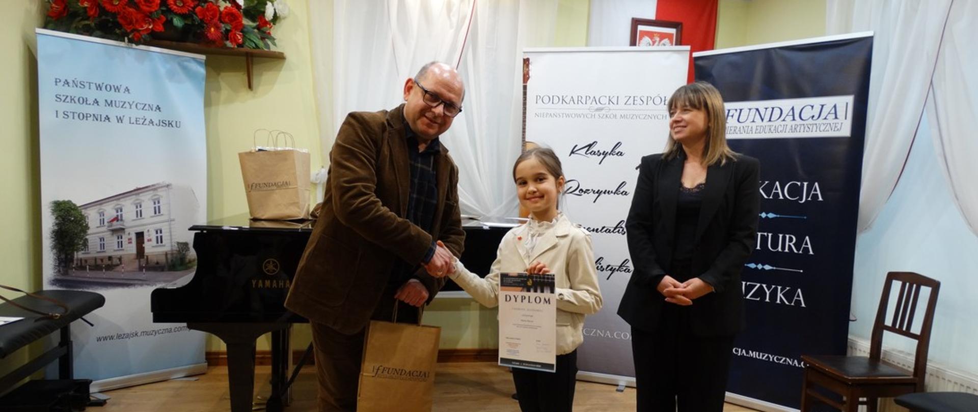 Organizator trzymający papierową torbę z prezentami konkursowymi gratuluje uczestniczce zdobytego miejsca. Uczennica prezentuje zdobyty dyplom, obok niej przedstawiciel organizatora. Za plecami osób ze zdjęcie widoczne rollupy organizatora