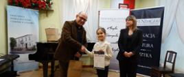Organizator trzymający papierową torbę z prezentami konkursowymi gratuluje uczestniczce zdobytego miejsca. Uczennica prezentuje zdobyty dyplom, obok niej przedstawiciel organizatora. Za plecami osób ze zdjęcie widoczne rollupy organizatora