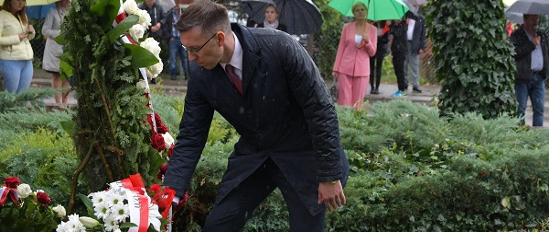 Na zdjęciu, na pierwszym planie elegancko ubranego mężczyznę w okularach, który składa wiązankę białych kwiatów z biało czerwoną wstążką. W tle widać kilka osób z parasolkami.