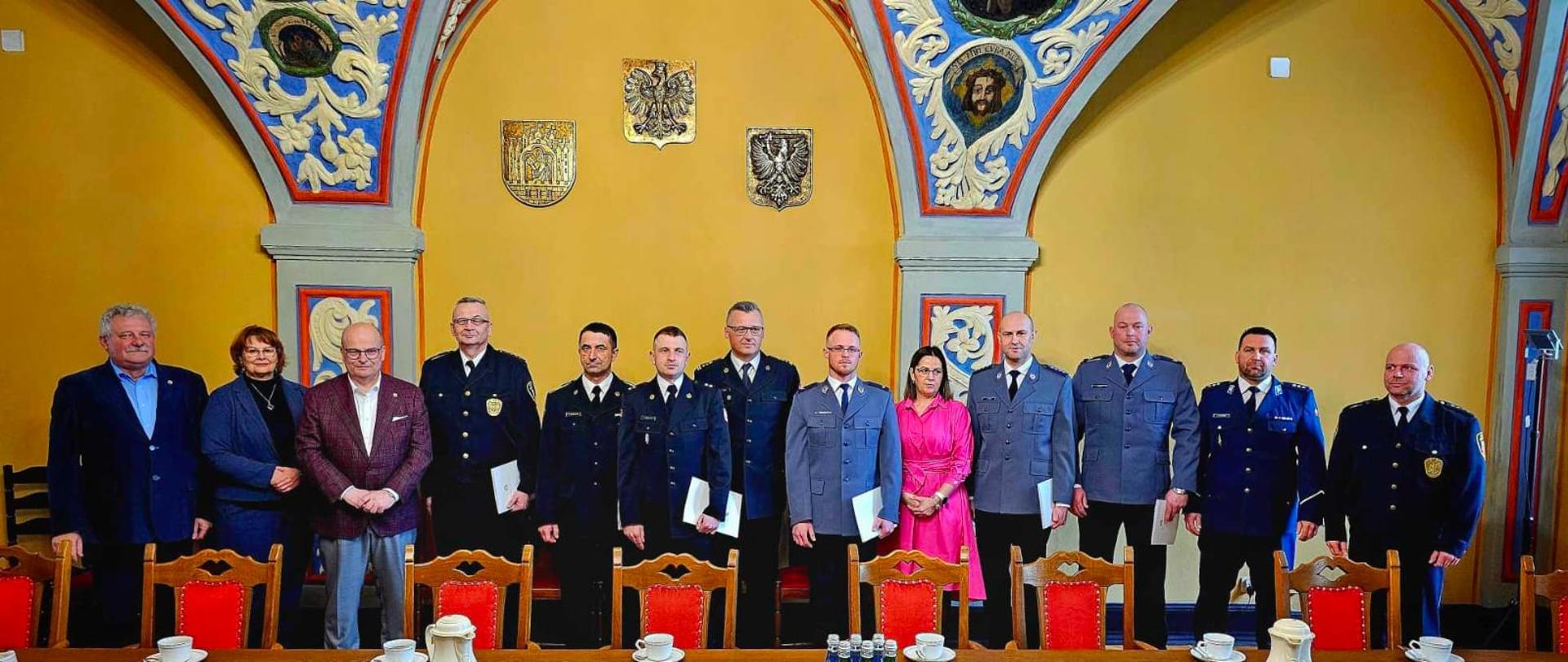 Zbiórka funkcjonariuszy w mundurach i osób cywilnych, które stoją w rzędzie w zabytkowym pomieszczeniu i pozują do zdjęcia.