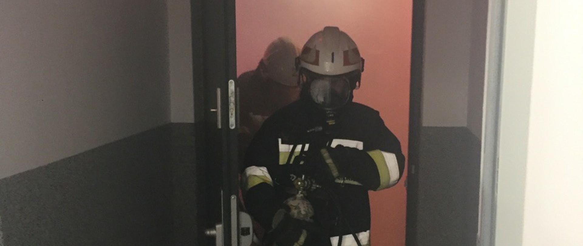 Zdjęcie przedstawia dwóch strażaków wychodzących z zadymionego pomieszczenia komory dymowej.