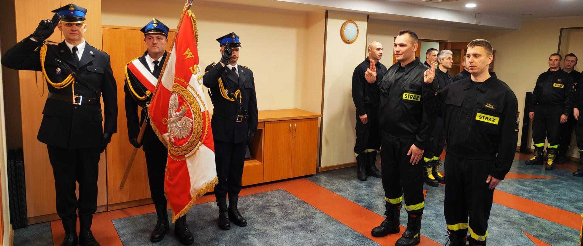 Zdjęcie zrobione w świetlicy remizy strażackiej. Z lewej strony stoi trzech strażaków w mundurach galowych ze sztandarem. Z prawej dwóch strażaków w mundurach koszarowych w trakcie ślubowania, uniesiona prawa ręka i złączone dwa palce. Za nimi reszta załogi.