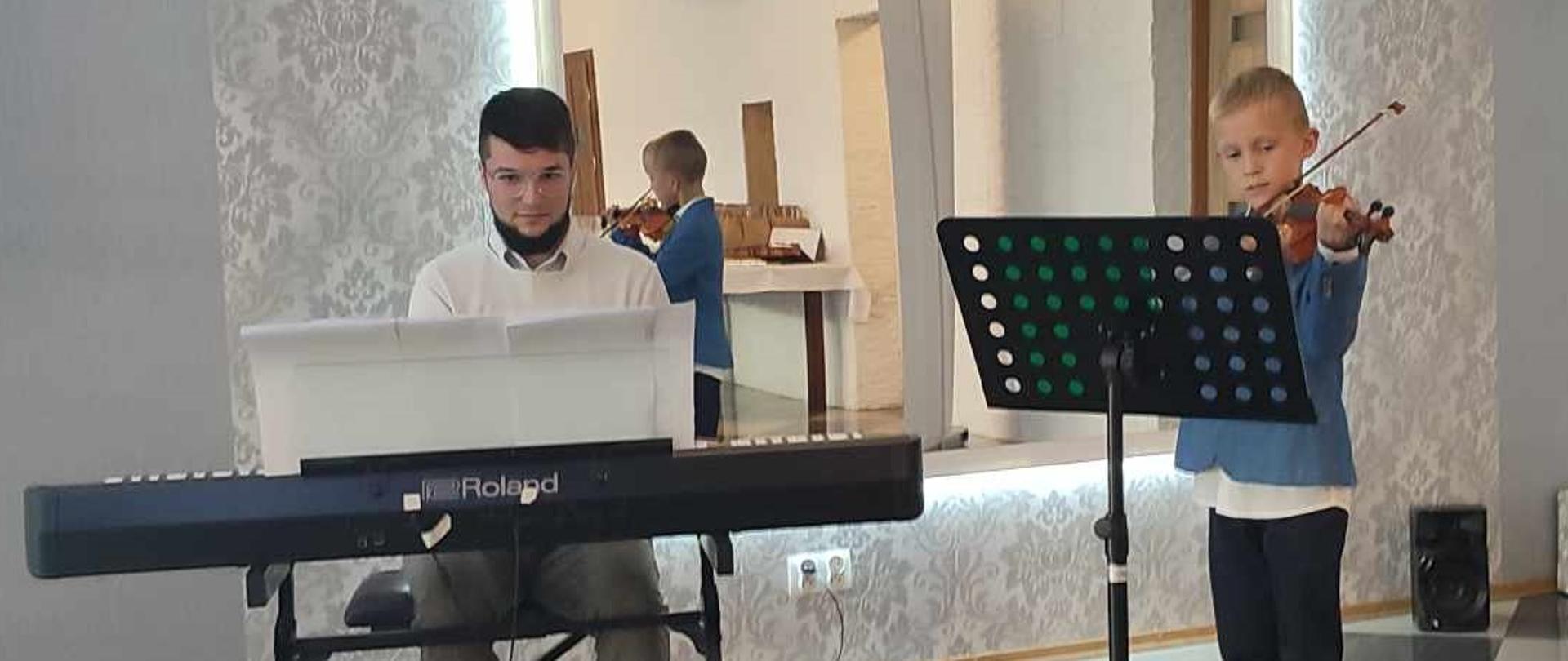 Chłopiec grający na skrzypcach i akompaniujący nauczyciel pan Krystian Klej. W tle wiszące bardzo duże lustro, w którym odbija się wizerunek chłopca.