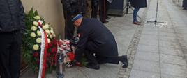 Na zdjęciu widzimy strażaka ubranego w płaszcz galowy koloru granatowego, rogatywka na głowie. Strażak składa kwiaty przed tablicą pamiątkową przymocowaną do ściany. Pod tablicą znajdują się także inne kwiaty i wieniec w biało czerwonych barwach. W tle widzimy osoby przyglądające się wydarzeniom. Dalej widzimy ulice oraz budynki mieszkalne. 