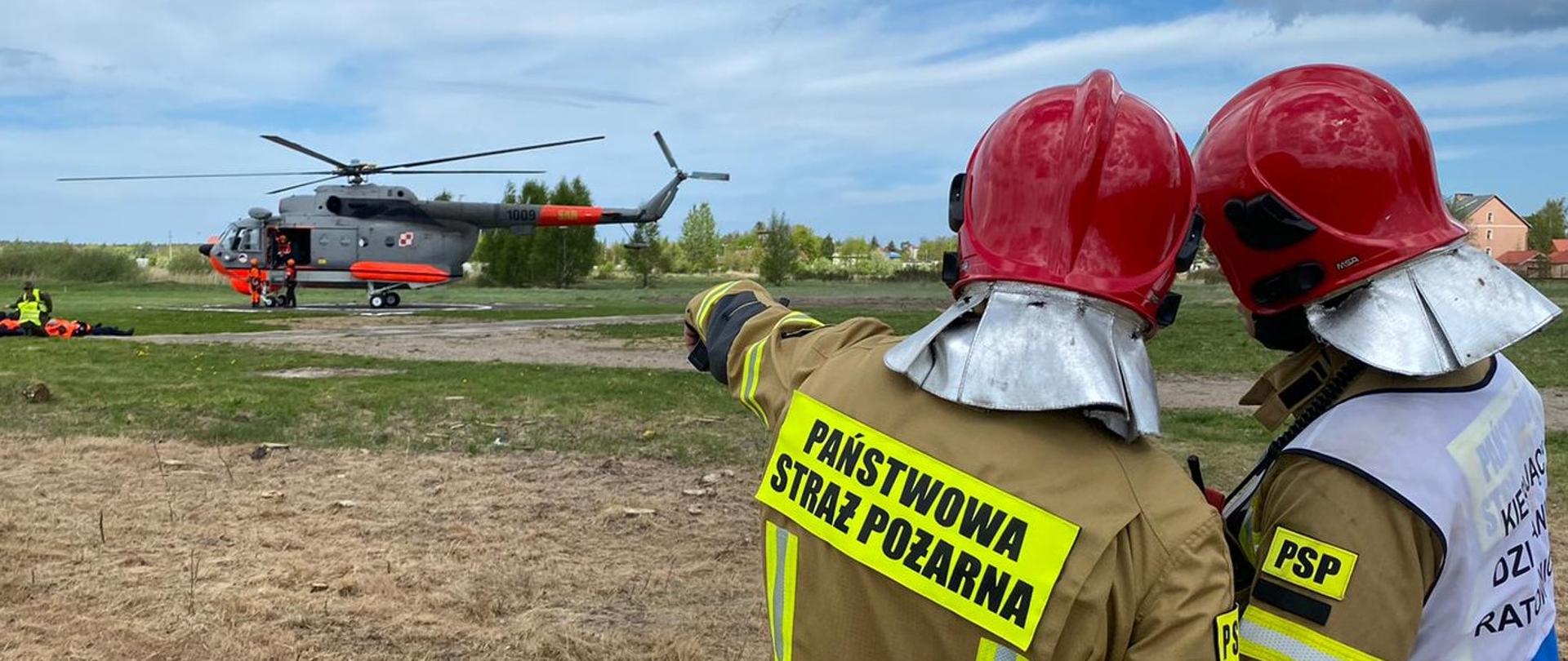 Na zdjęciu dwóch strażaków wskazuje na helikopter na lądowisku