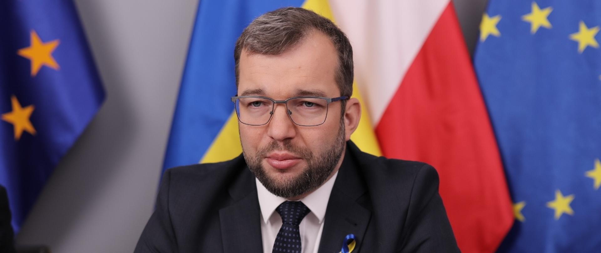 Minister Grzegorz Puda przy biurku, w tle flagi Polski, Ukrainy i Unii Europejskiej