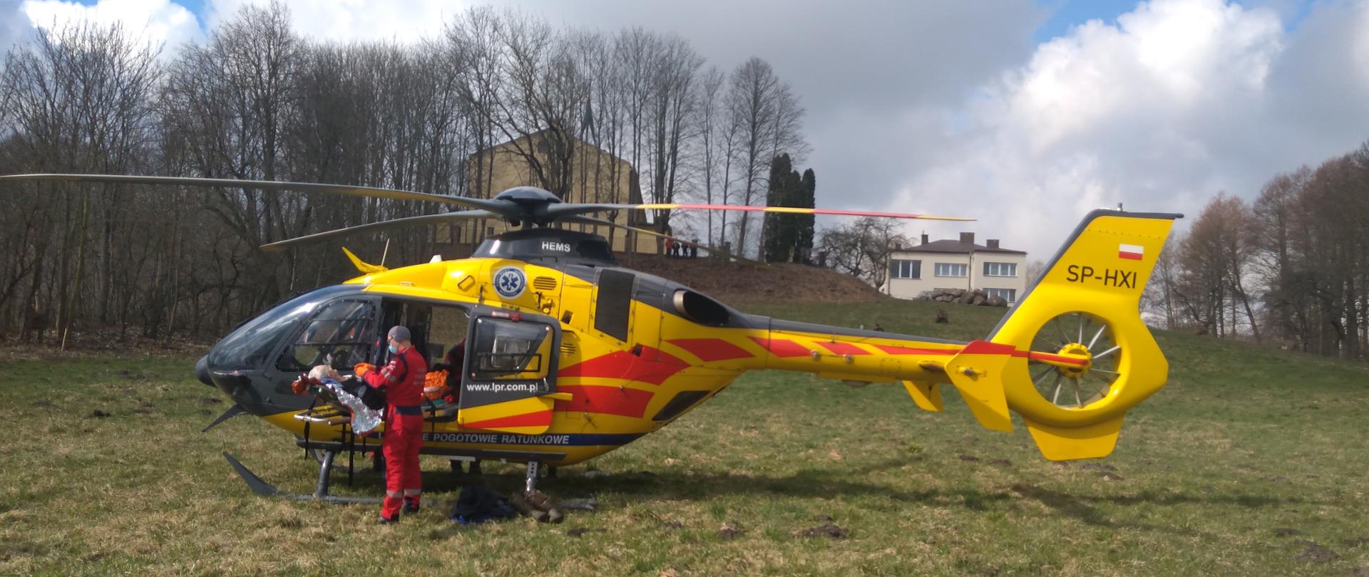 Ratownik Lotniczego Pogotowia Ratunkowego udziela pomocy medycznej mężczyźnie. Poszkodowany leży na noszach przy śmigłowcu. Zdarzenie ma miejsce na łące z dala od zabudowań. 