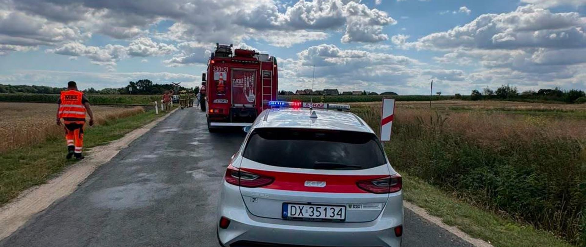 Samochód operacyjny i wóz strażacki zabezpieczają miejsce działań ratowniczych na wiejskiej drodze. W tle strażacy prowadzą akcję, a po obu stronach drogi znajdują się pola uprawne.