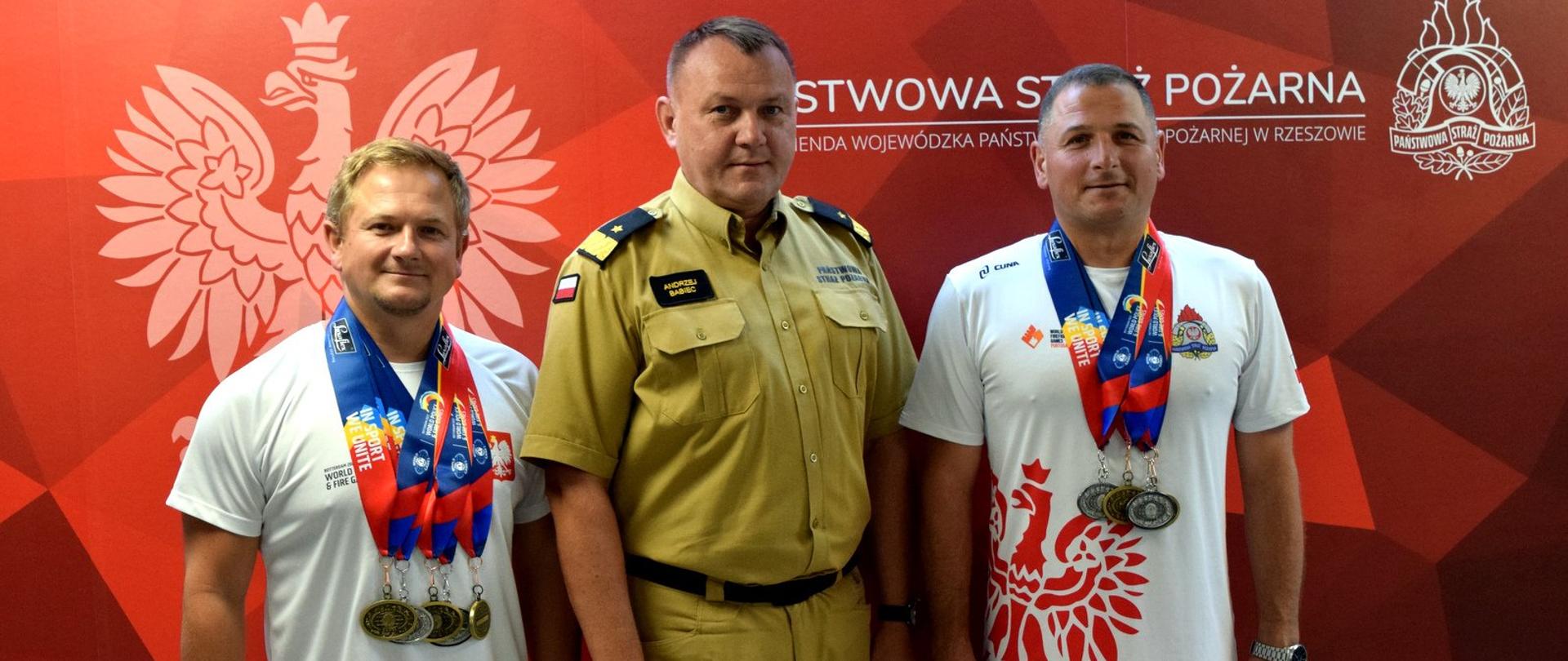 Zdjęcie zrobione w pomieszczeniu. W linii stoją trzy osoby. Po lewej i prawej stronie mężczyźni w strojach sportowych z medalami. W środku stoi oficer PSP w stopniu nadbrygadiera. 