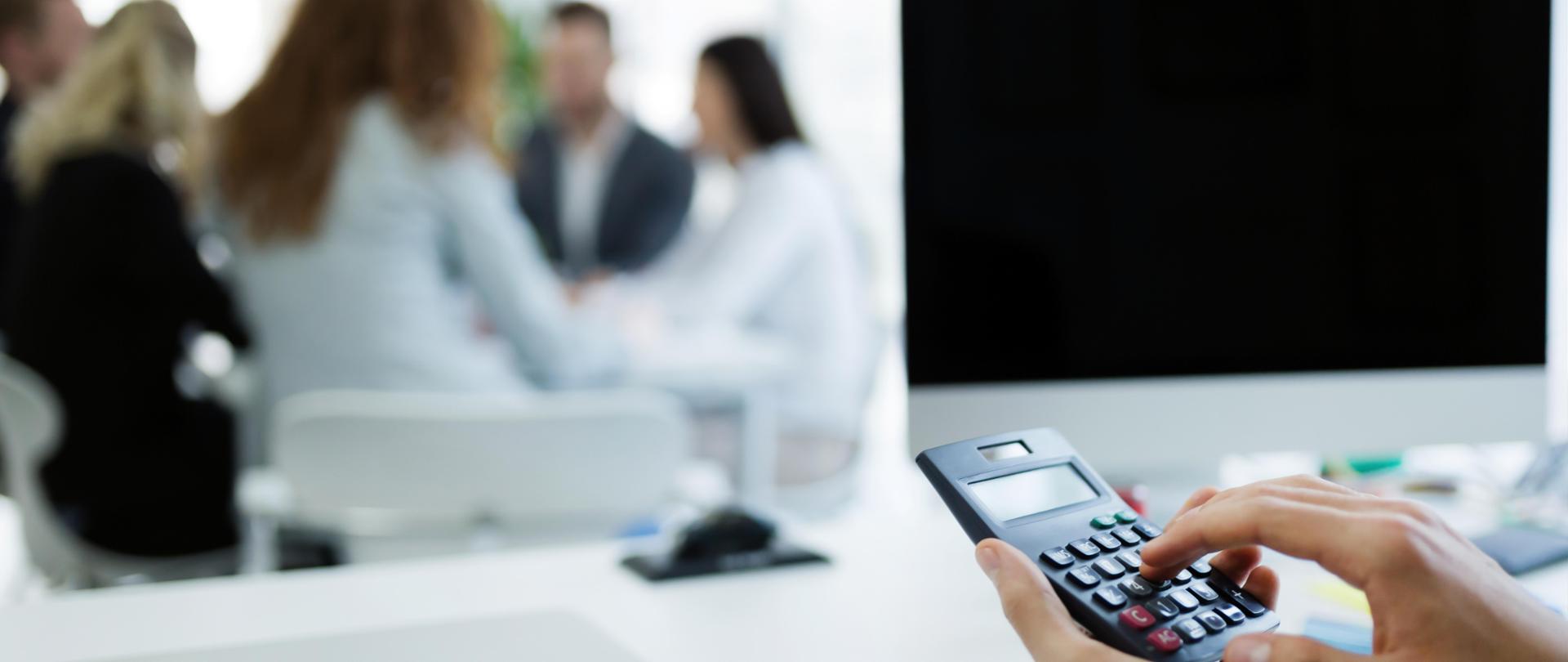 Na pierwszym planie kalkulator a w tle grupa dyskutujących osób