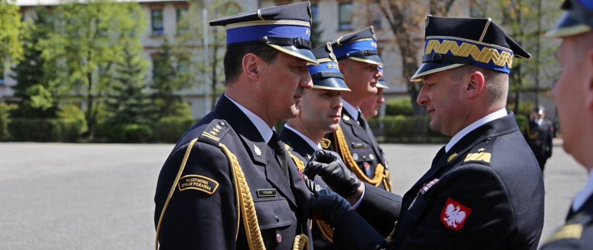 Zastępca komendanta głównego PSP przypina odznaczenie komendantowi szkoły, za którym stoją w szeregu inni strażacy