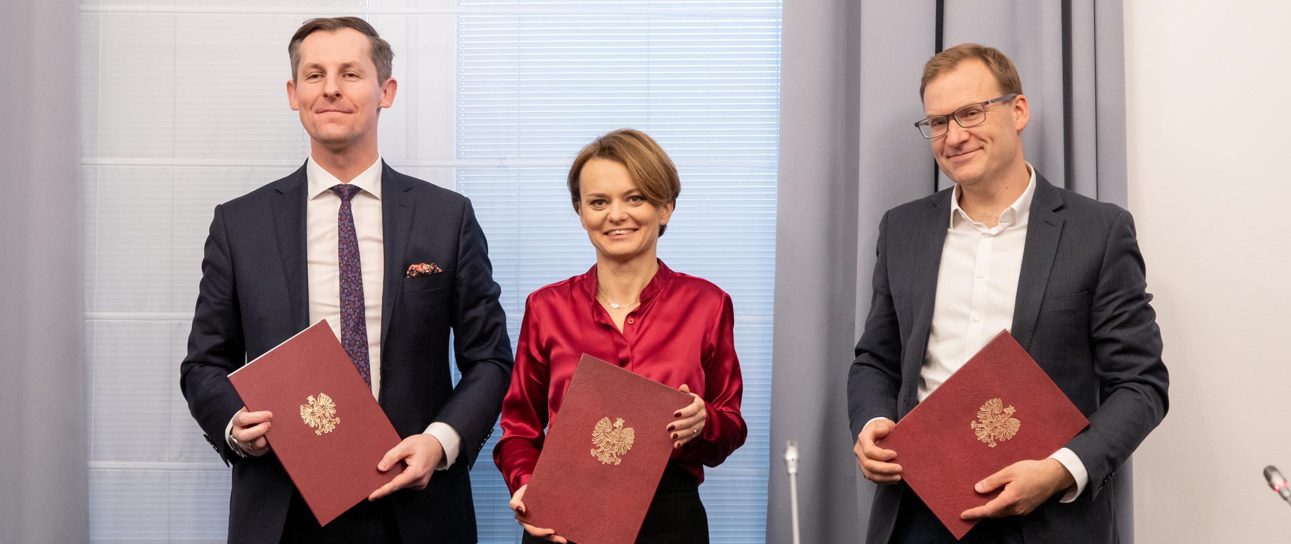 Od lewej stoją wiceprezes UOKiK, minister rozwoju, przedstawiciel Polskiego Alarmu Smogowego. W rekach trzymają teczki z podpisanych porozumieniem.